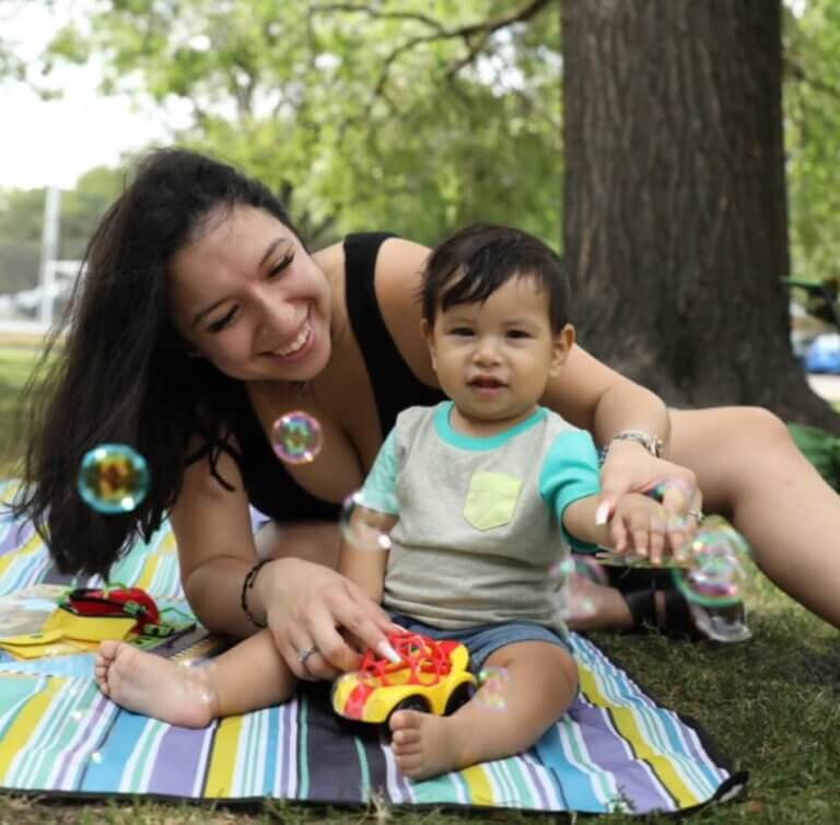 A baby and mother sitting on a picnic blanket in a park as bubbles float by.