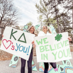 2 Kappa Delta sisters holding signs that read "Kappa delta loves you" and "We believe in you"