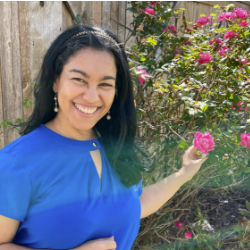 A woman smiling, standing in front of a dark pink rose bush, wearing a deep blue blouse.