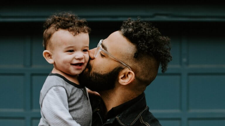 A man, with clear glasses and curly dark hair, kisses the cheek of a smiling baby, with curly brown hair, that he is holding in his arms.