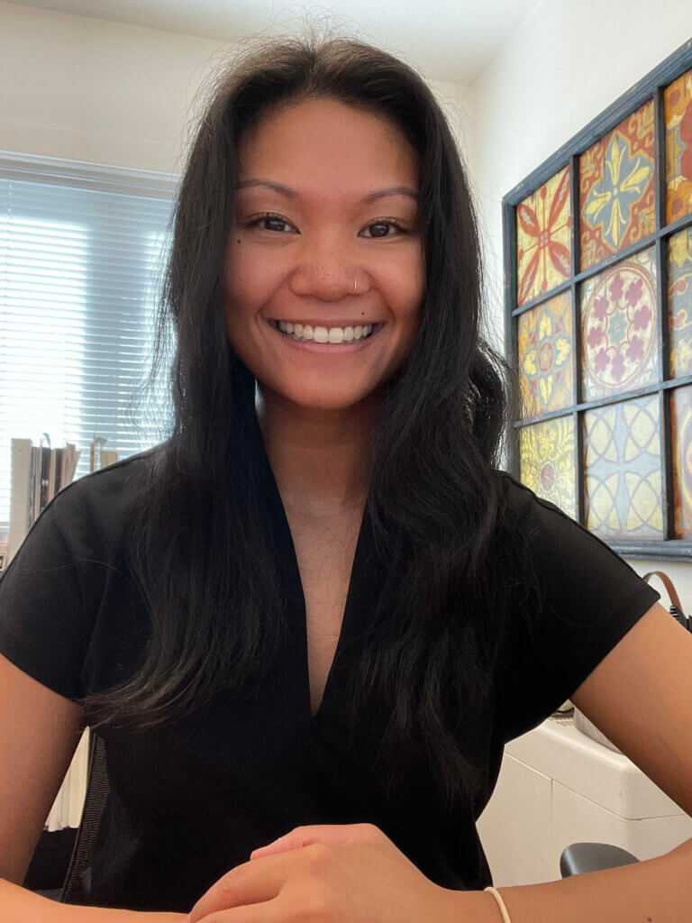 A photo of Jade Angulo sitting at a desk. She is a wearing a v-neck black shirt and smiling.