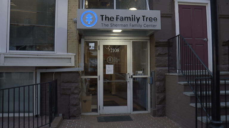 A photo of a glass door at night, with a sign that says "The Family Tree: The Sherman Family Center" with a blue neon sign in the shape of a circle with two overlapping arrows pointing upward.